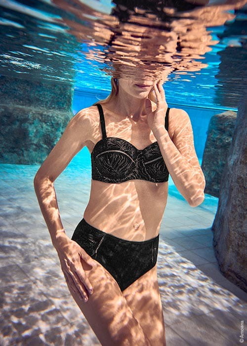 Unterwasseransicht einer Frau in schwarzem Prothesen-Bikini, die in einem Pool mit Naturstein-Elementen steht.