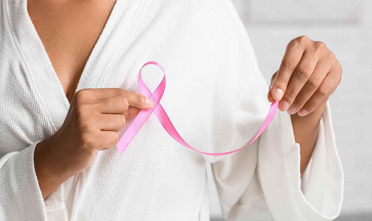 Oberkörper einer Frau im weißen Bademantel, die eine rosa Brustkrebs-Schleife vor ihre linke Brust hält