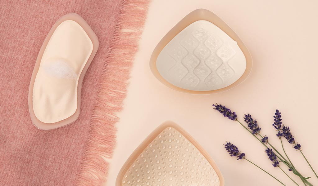 Drei unterschiedliche Brustprothesen, die auf einem rosanen Tuch und pastellfarbenem Untergrund liegen