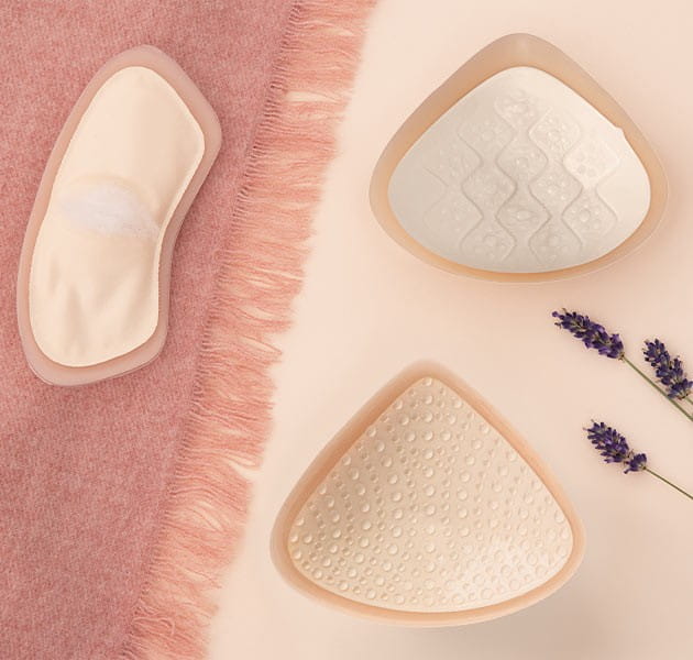 Drei unterschiedliche Brustprothesen, die auf einem rosanen Tuch und pastellfarbenem Untergrund liegen