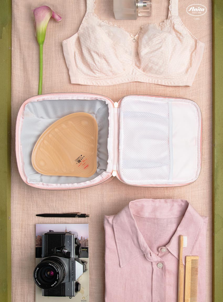 Verschiedene Dinge, packfertig bereitgelegt für die Reise: Ein Prothesen-BH, ein Oberteil, Zahnbürste, Kamera und Stift. In der Mitte steht prominent ein offenes Prothesen-Etui, in dem eine Anita Brustprothese liegt.
