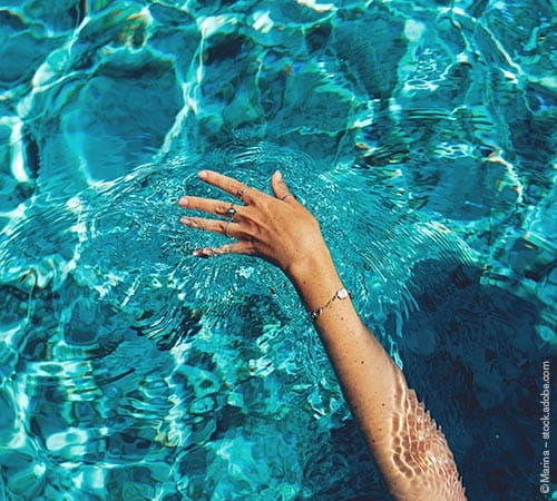 Der Arm einer Frau, geschmückt mit Ringen und Armband, der sanft über die Wasseroberfläche eines blauen Swimmingpools gezogen wird.