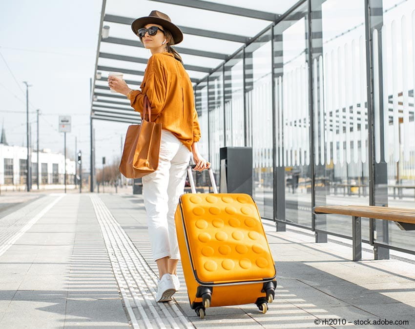 Eine Frau in legerer Kleidung mit Sonnebrille, Hut und Reisegepäck an einer Haltestelle.